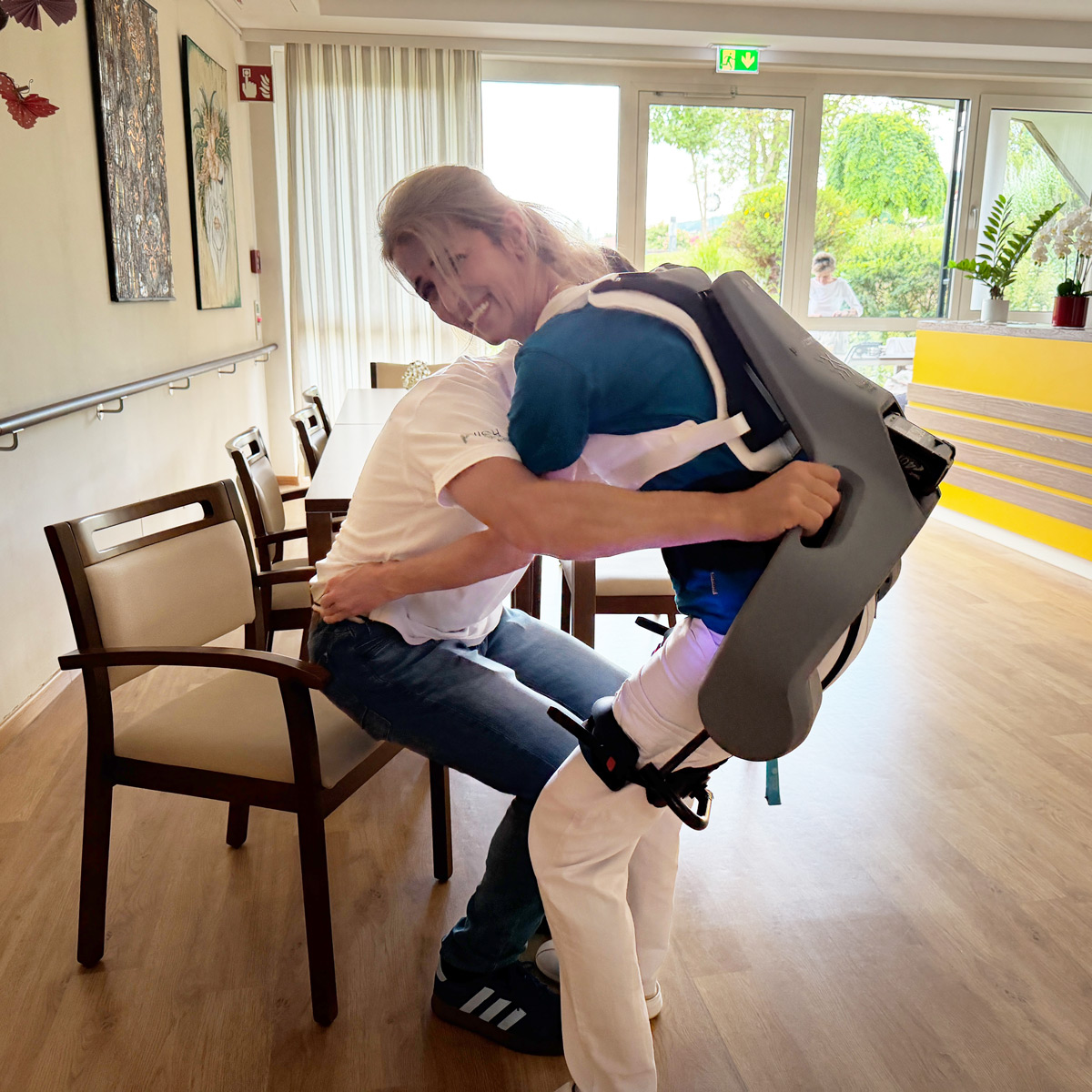 Pflegekraft hebt Person aus dem Sitzen mit Hilfe des Exoskeletts