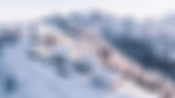 A snow-covered mountain village with white houses, a church, and frosty trees, framed by a backdrop of rugged, snowy peaks under a clear sky.