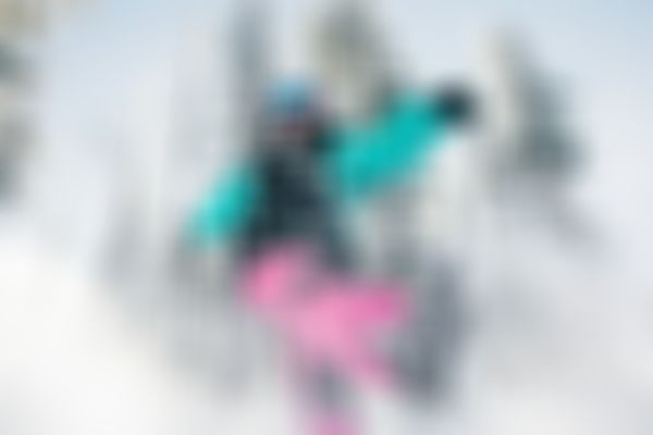 Person in vibrant snowboard gear kicking up snow, surrounded by snow-covered trees under a clear blue sky.