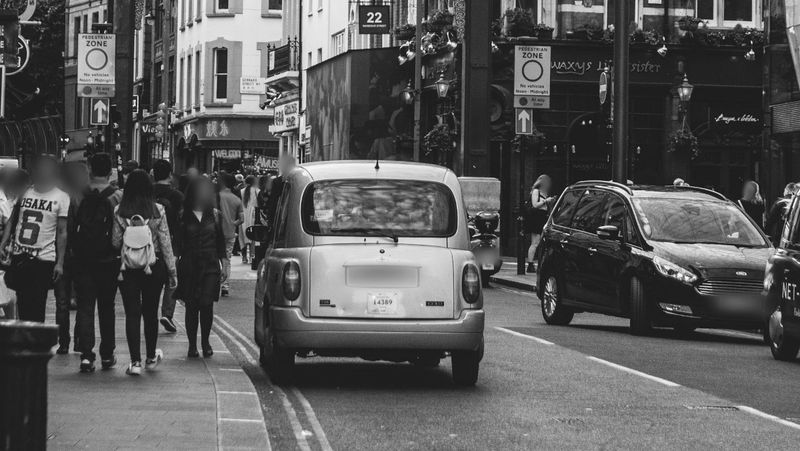 Black and white photo of a busy city street with cars and pedestrians walking along the sidewalk.