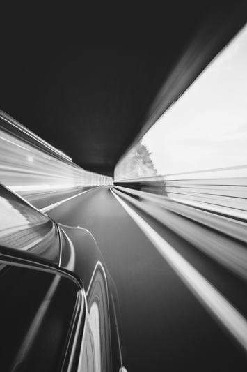 Imagen en blanco y negro de un automóvil que circula a toda velocidad por un túnel, lo que crea un desenfoque de movimiento y líneas dinámicas en la carretera y las paredes del túnel.