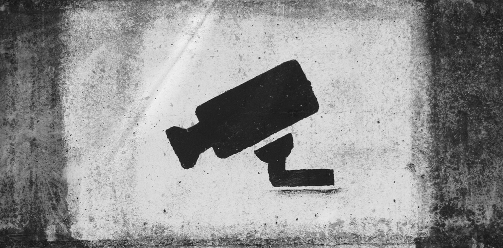 Graffiti przedstawiające czarną kamerę monitorującą na zniszczonej, teksturowanej ścianie tworzy ostry, miejski kontrast.