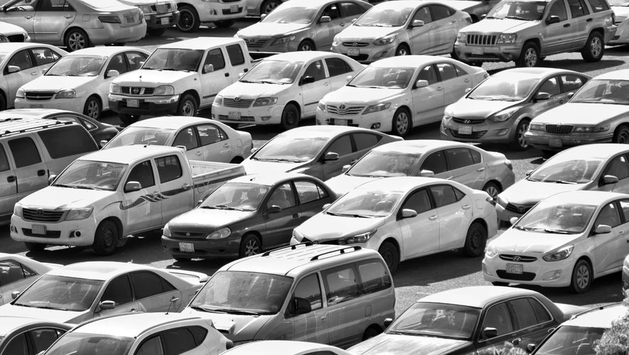 Zatłoczony parking wypełniony różnymi samochodami, w tym sedanami, SUV-ami i minivanami, wszystkie zaparkowane blisko siebie. Obraz jest czarno-biały.