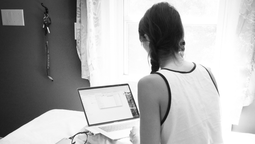 Woman with braided hair using a laptop in a bright room, sitting on a bed near a window with keys hanging on the wall.