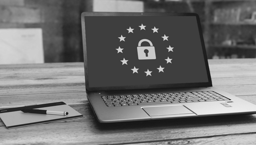 Laptop mit Schlosssymbol, umgeben von Sternen, symbolisiert Datenschutz, auf einem Holztisch. Daneben ein Stift und ein Notizblock. Schwarzweiß.