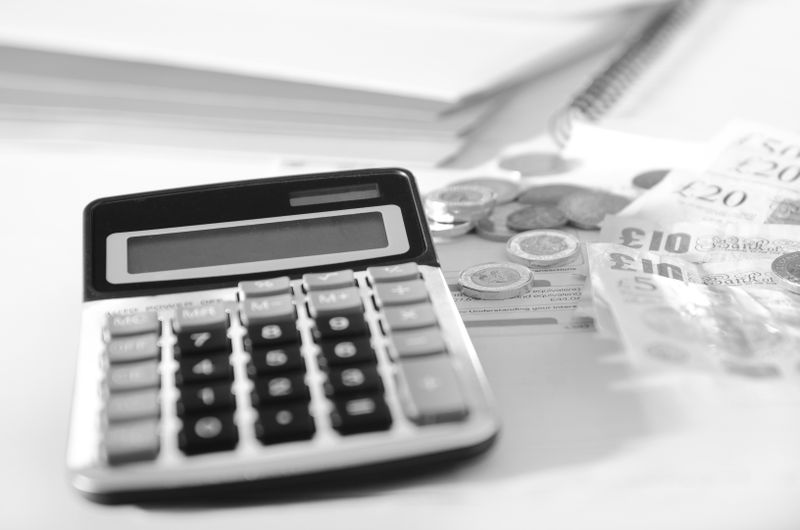 Calculadora con billetes y monedas sobre un escritorio, junto a un cuaderno de espiral y papeles. Imagen en blanco y negro.