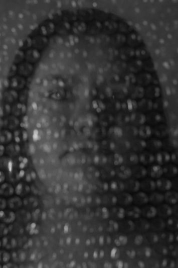 A person's face is visible through a layer of bubble wrap, creating a textured and abstract effect in grayscale.