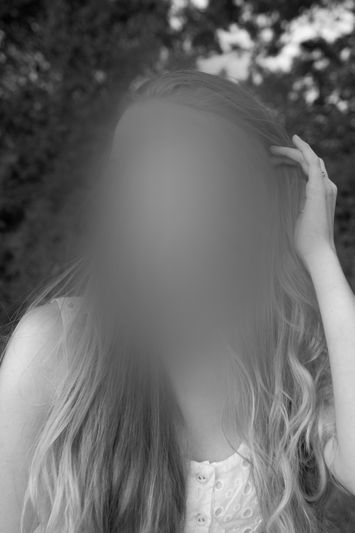 Czarno-białe zdjęcie portretowe osoby z długimi włosami na zewnątrz; twarz rozmyta, ręka dotyka włosów, ubrana w zapinaną na guziki koszulkę bez rękawów.