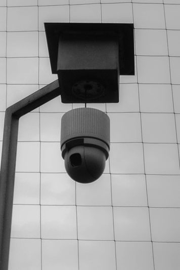Black and white image of a security camera mounted on a pole, set against a tiled wall background.