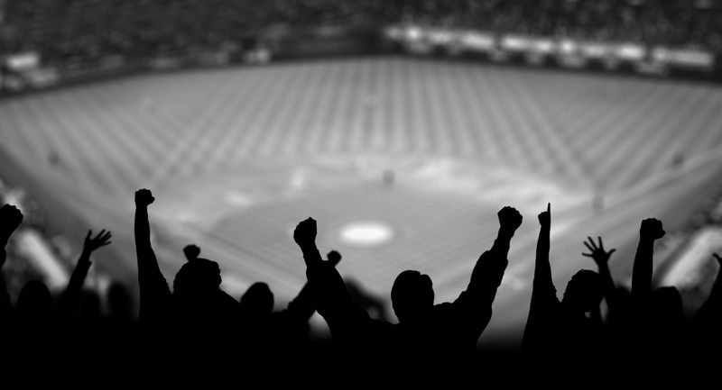Siluetas de aficionados animando en un estadio con una vista borrosa de un campo de béisbol en el fondo.