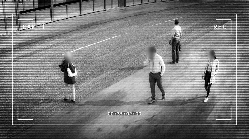 Imagen en blanco y negro de una cámara de vigilancia urbana que muestra a peatones con las caras difuminadas junto a la carretera