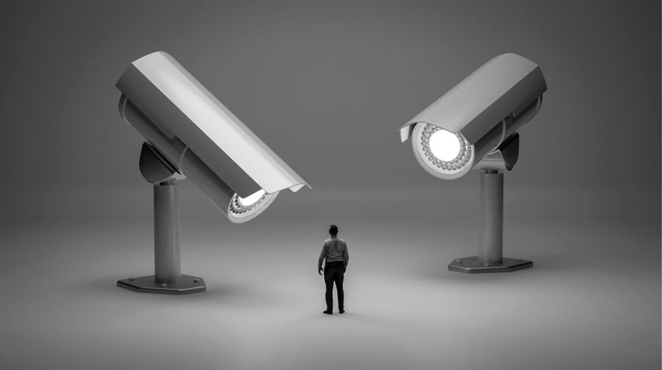 A person stands between two large surveillance cameras facing each other in a monochromatic setting.