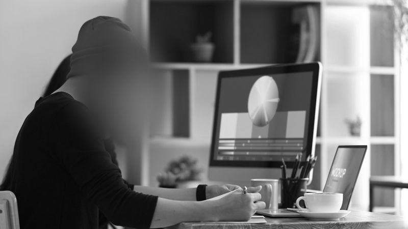 Personne portant un bonnet travaillant à un bureau avec un ordinateur et un ordinateur portable, tasses de café à proximité, dans un espace de bureau minimaliste.