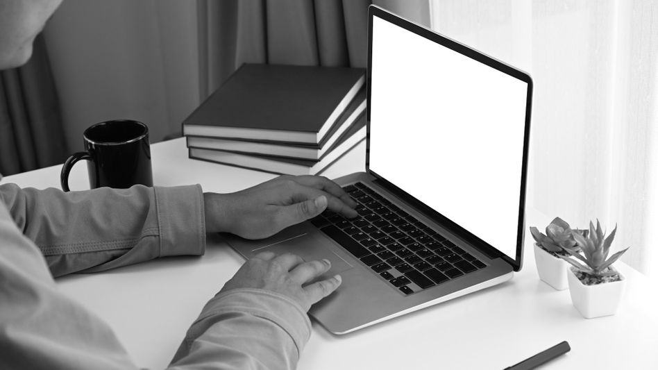 Une personne tape sur un ordinateur portable à un bureau blanc avec des livres empilés, une tasse et une petite plante en pot à proximité.