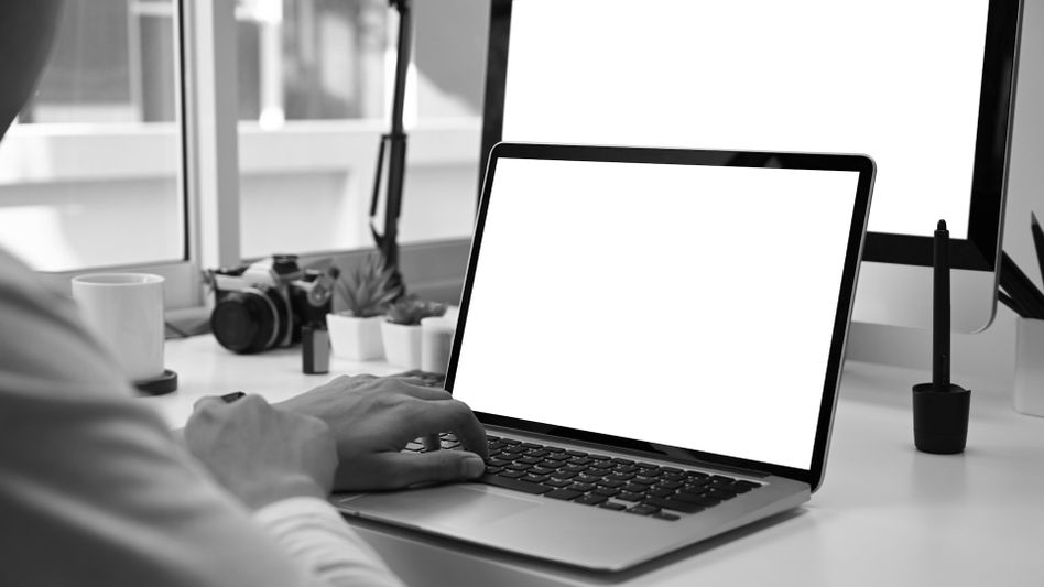 Une personne utilise un ordinateur portable avec un écran vide à son bureau, entourée d'une caméra, de plantes et d'un grand écran. Image en noir et blanc.