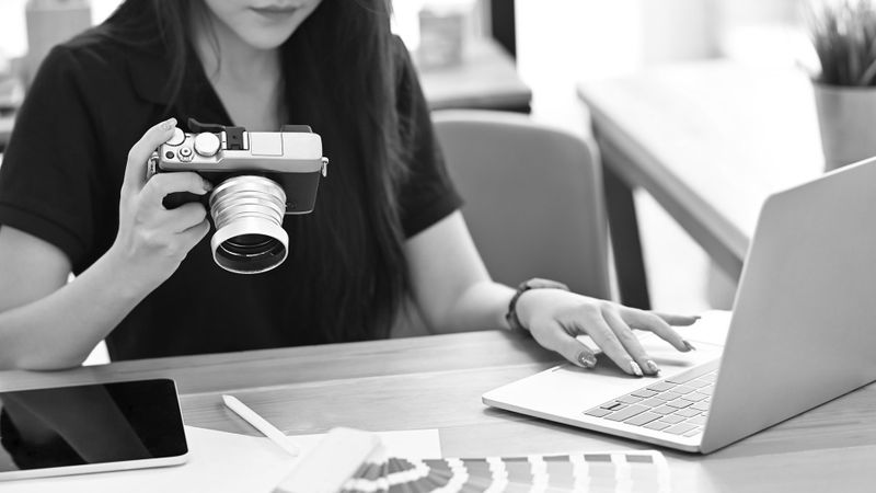 Person holding a camera and using a laptop at a desk, with a tablet and color swatches nearby.