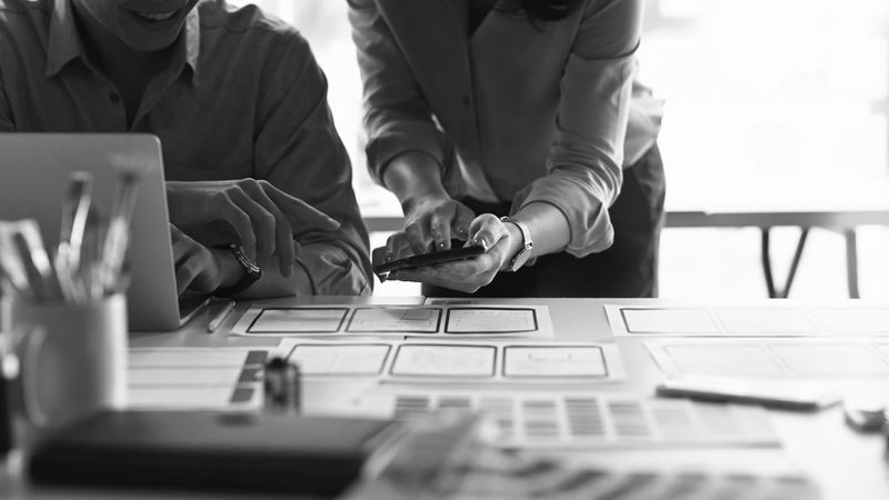 Deux personnes collaborant à un bureau, discutant de documents et d'un smartphone. La scène transmet le travail d'équipe et la planification. Image en noir et blanc.
