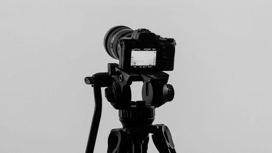 Black camera on a tripod facing away, with a visible LCD screen displaying settings, against a plain gray background.