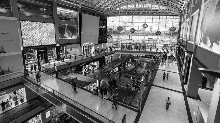 Czarno-biały szeroki widok na atrium wielopoziomowego centrum handlowego z ruchomymi schodami, szklanym dachem, sklepami i klientami.