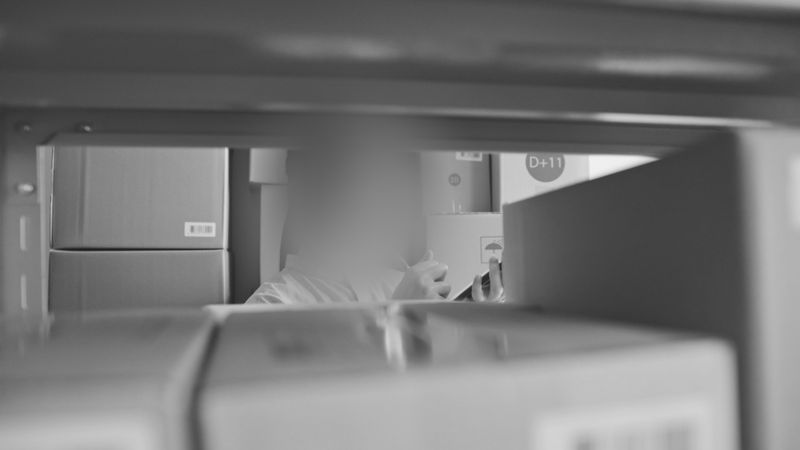 Vue à travers des étagères remplies de cartons empilés, avec une silhouette floue en arrière-plan en train de ranger des articles.