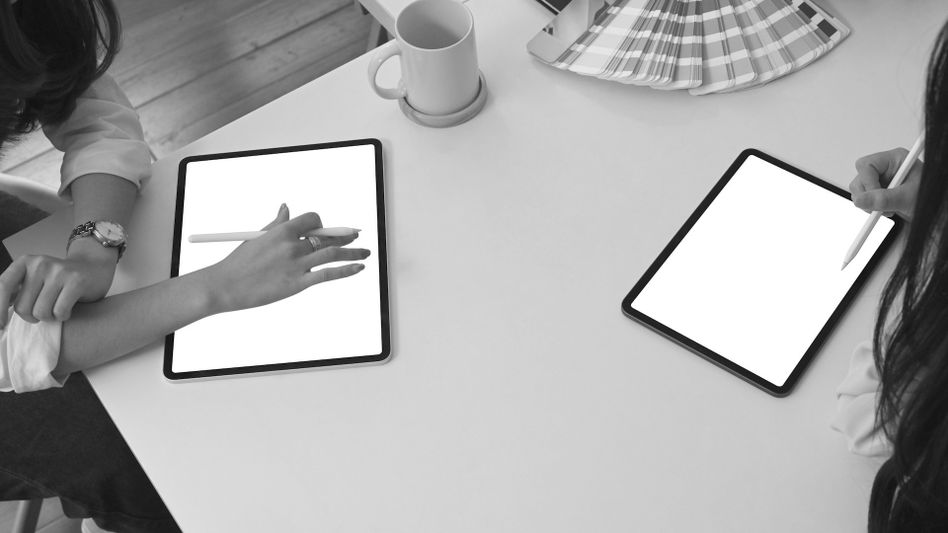 Deux personnes utilisent des stylets sur des tablettes, assises à une table blanche, avec une tasse à café et des échantillons de couleurs à proximité. Image en noir et blanc.