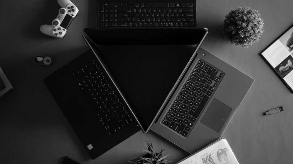 Trois ordinateurs portables forment un triangle sur une table, avec une manette de jeu, une plante, un carnet et un magazine à proximité. Image en noir et blanc.