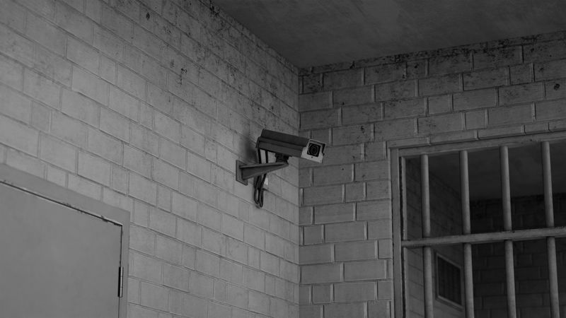 a surveillance camera in a prison attached to a white brick wall, with bars visible in the background