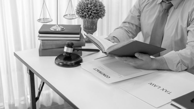 Personne lisant un livre à un bureau avec des documents juridiques, un marteau de juge, des balances et une plante en pot à proximité.