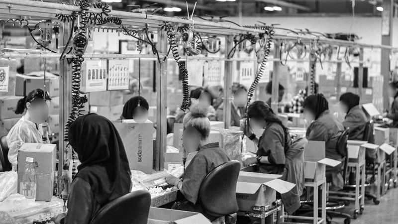 L'importance de l'anonymisation des vidéos dans les services de santé et de sécurité Des ouvriers sur une chaîne de montage d'usine, assis et concentrés sur leurs tâches, entourés de boîtes et d'équipements, dans un environnement industriel animé.