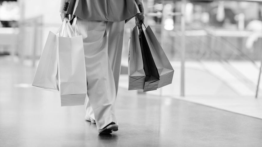 Osoba idąca po centrum handlowym, niosąca w obu rękach kilka toreb z zakupami. Czarno-białe zdjęcie skupia się na dolnej części ciała.