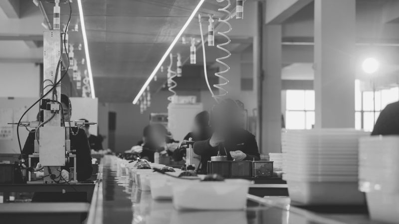 Monochrome image of a factory assembly line with workers handling items, fluorescent lights overhead, and trays on the conveyor belt.