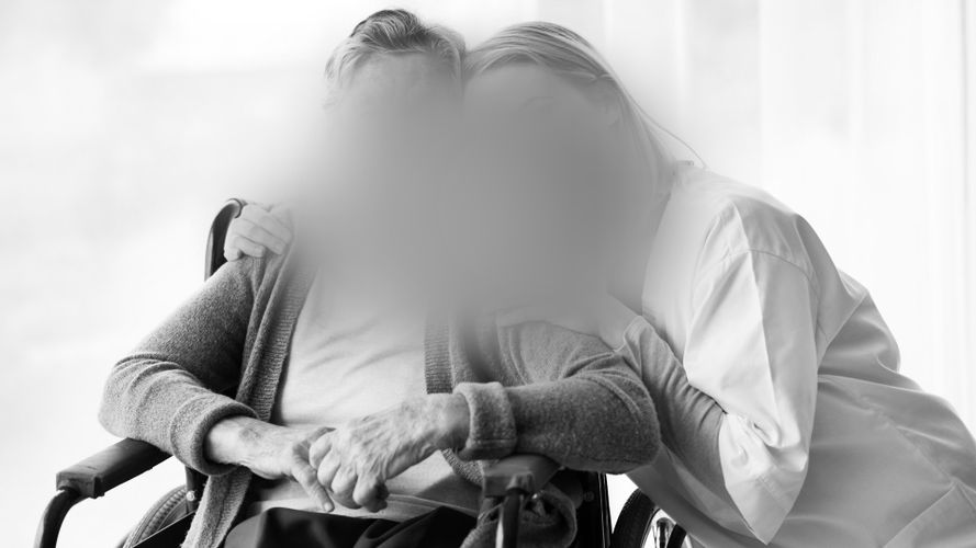 Una imagen en blanco y negro de dos personas con los rostros borrosos; una está sentada en una silla de ruedas y la otra se inclina abrazando a la persona sentada.