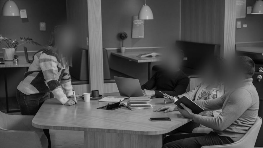 Cztery osoby siedzą przy dużym drewnianym stole, mając przed sobą otwarte laptopy i zeszyty. Jedna z nich stoi, a pozostałe siedzą i prowadzą rozmowę.