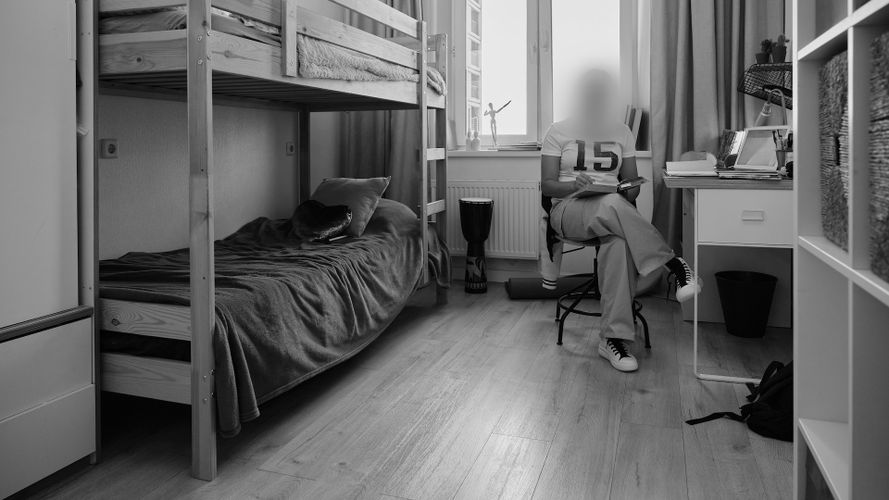 Una persona se sienta en una silla junto a un escritorio en una habitación ordenada con una litera, ventana y suelo de madera.