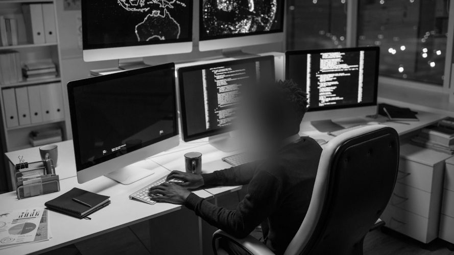 Eine Person sitzt an einem Schreibtisch mit mehreren Monitoren, die Code und Karten anzeigen, arbeitet in einem schwach beleuchteten Büro, das Gesicht unscharf, und eine Tasse steht in der Nähe.
