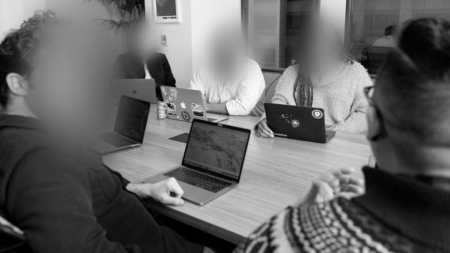 Eine Gruppe von Menschen in einem Besprechungsraum, die um einen Tisch mit Laptops sitzen und eine Diskussion führen. Ihre Gesichter sind verschwommen.