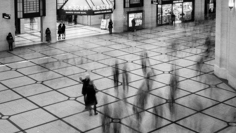 Fotografía en blanco y negro de una concurrida plaza interior con figuras borrosas caminando, piso de baldosas y carteles iluminados en el fondo.