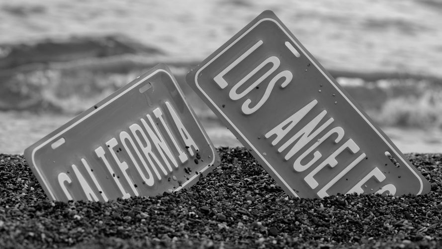 Czarno-białe zdjęcie tablic rejestracyjnych z Kalifornii i Los Angeles częściowo zakopanych w kamykach na plaży, z falami w tle.