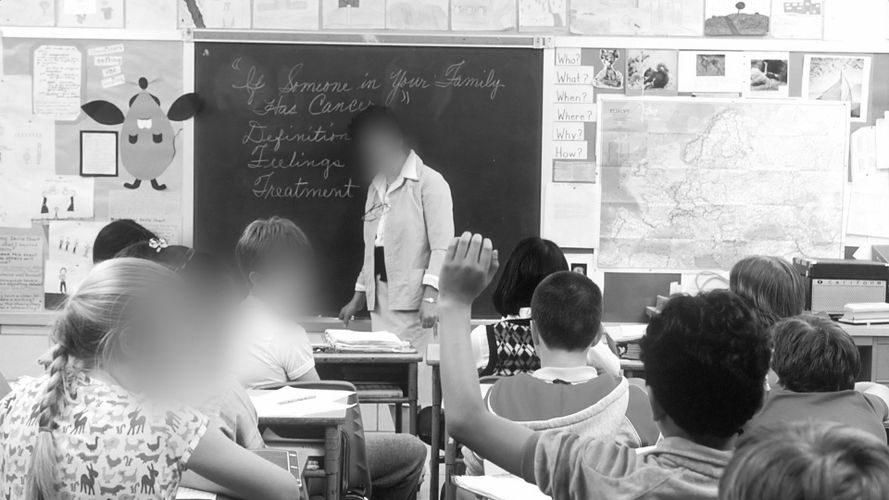A classroom with students seated, one raising a hand, and a teacher standing at the blackboard. Maps and posters decorate the walls.