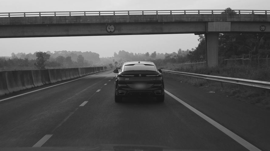Czarno-białe zdjęcie samochodu jadącego autostradą, zbliżającego się do wiaduktu, z drzewami i barierami wzdłuż drogi.