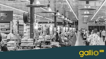 Czarno-białe zdjęcie ruchliwego supermarketu z wieloma kasami i kupującymi. Światła sufitowe odbijają się od błyszczącej podłogi.