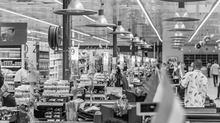 Czarno-białe zdjęcie tętniącej życiem okolicy przy kasach w supermarkecie, z ludźmi, wózkami na zakupy i wystawionymi produktami.