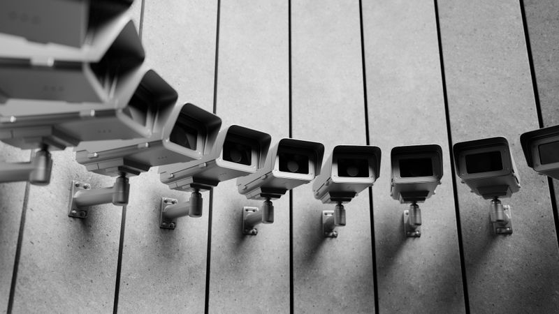 Fila de cámaras de vigilancia montadas en una pared, todas orientadas en la misma dirección, creando un patrón simétrico. Imagen en blanco y negro.