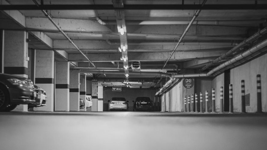 Czarno-białe zdjęcie podziemnego parkingu z zaparkowanymi samochodami, pasami na filarach i oświetleniem sufitowym.