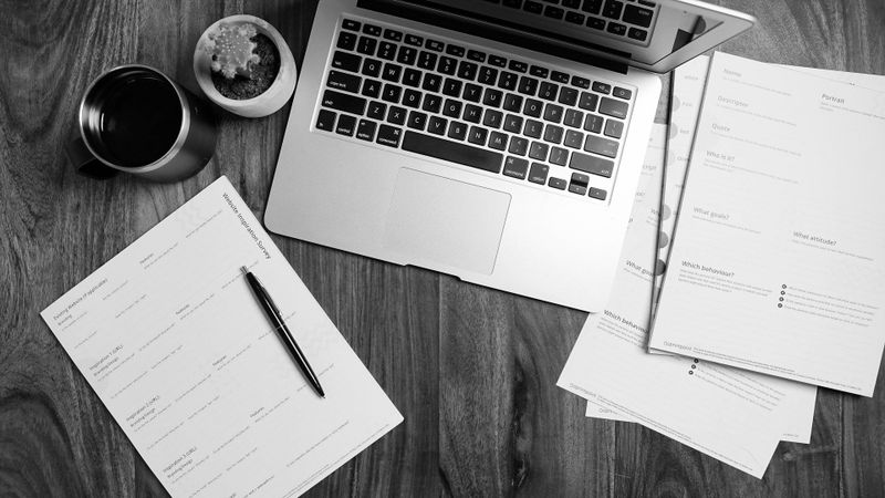 A laptop on a wooden desk, surrounded by a cup of coffee, documents, and a pen.