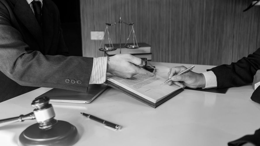 Dwie osoby w garniturach wymieniają się clipboardem i długopisem w biurze, w tle znajdują się wagi prawnicze, młotek sędziego i książki. Zdjęcie czarno-białe.