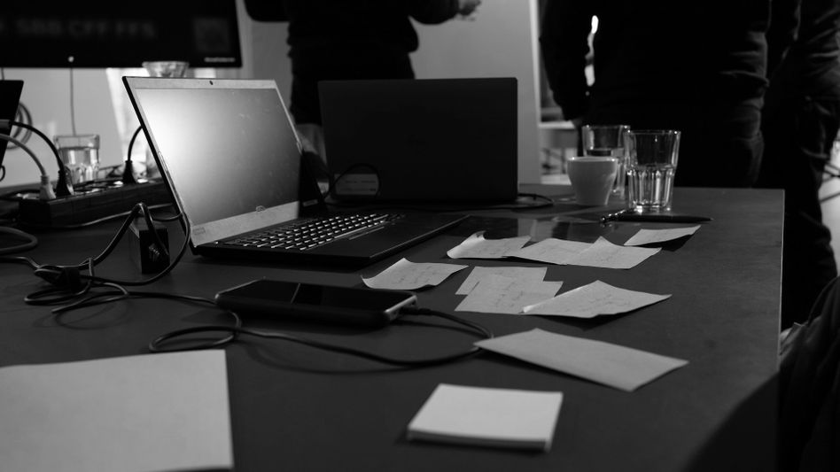 Imagen en blanco y negro de un espacio de trabajo con una laptop, un teléfono inteligente, notas adhesivas y vasos de agua sobre una mesa. Personas de pie al fondo.
