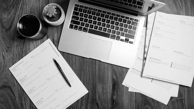 Una laptop sobre un escritorio de madera rodeada de documentos dispersos, una taza de café y un bolígrafo sobre un planificador.