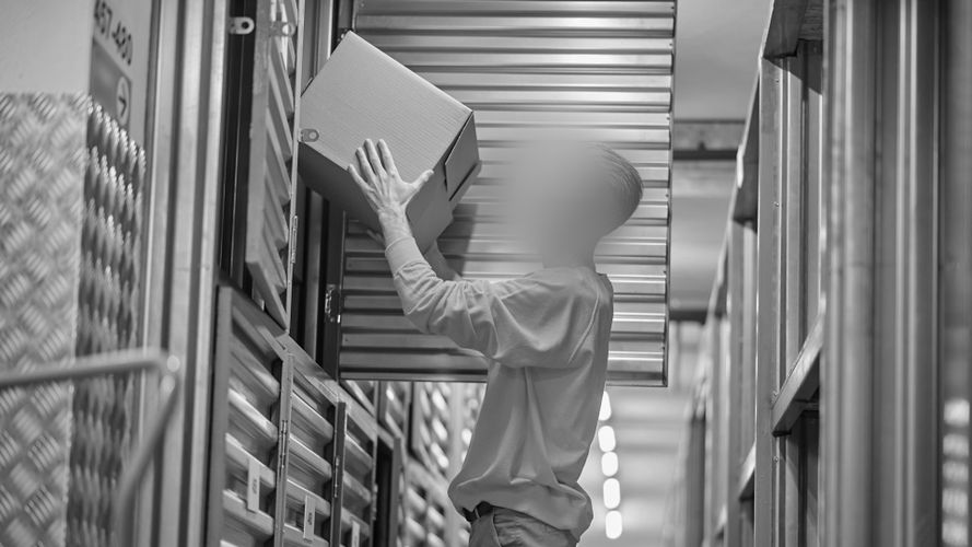 Osoba wkładająca pudełko do jednostki magazynowej z drzwiami z falistego metalu, w korytarzu z metalowymi ścianami i oświetleniem sufitowym.