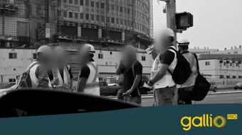 Varios trabajadores de la construcción con chalecos y cascos conversan en un sitio de construcción. Un edificio grande es parcialmente visible al fondo.